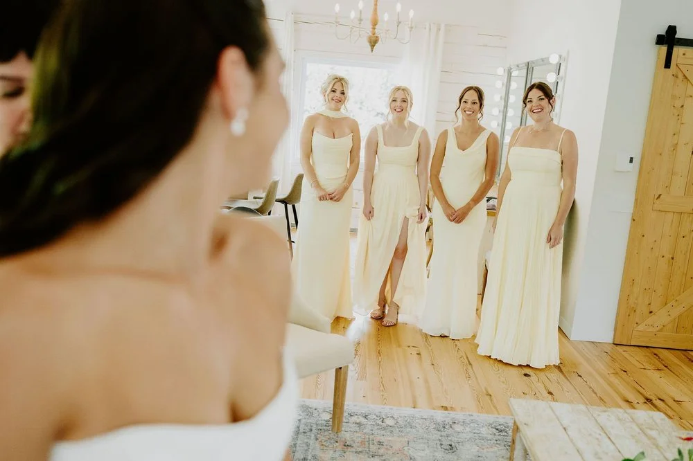 Bridesmaids in pale yellow gowns smiling as they see Kylie in her wedding dress for the first time