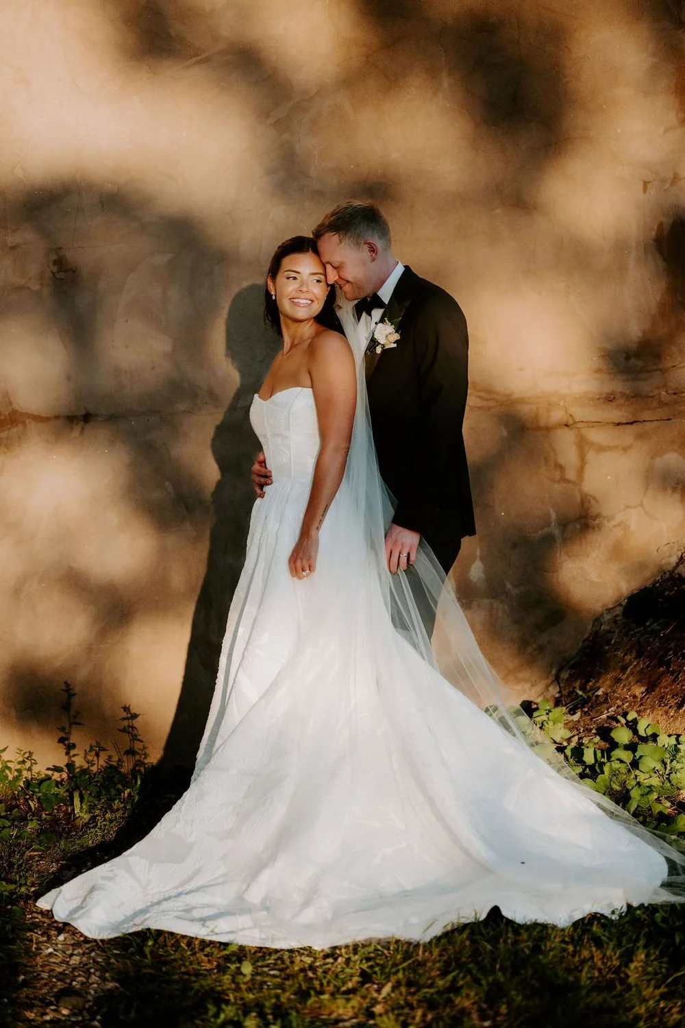 Kylie and Will embracing against a wall dappled with leaf shadows in the evening light