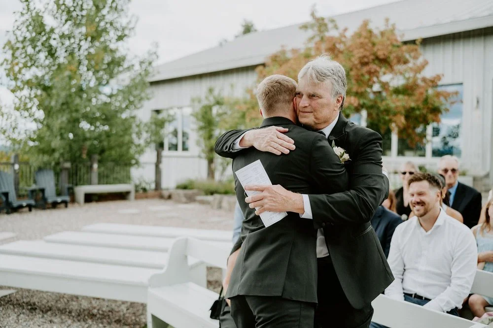 An older man in a tuxedo hugging Will beside the ceremony benches before the wedding