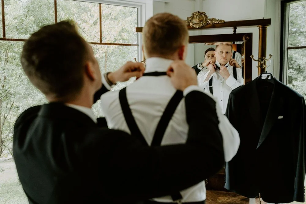 A groomsman fastening Will's bow tie as he watches in an ornate gilded mirror