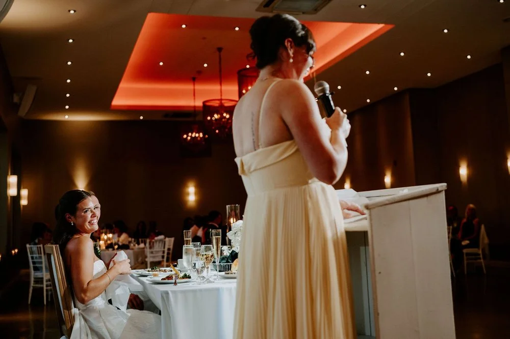 Kylie laughing at the head table as a bridesmaid in a pale yellow gown gives her speech