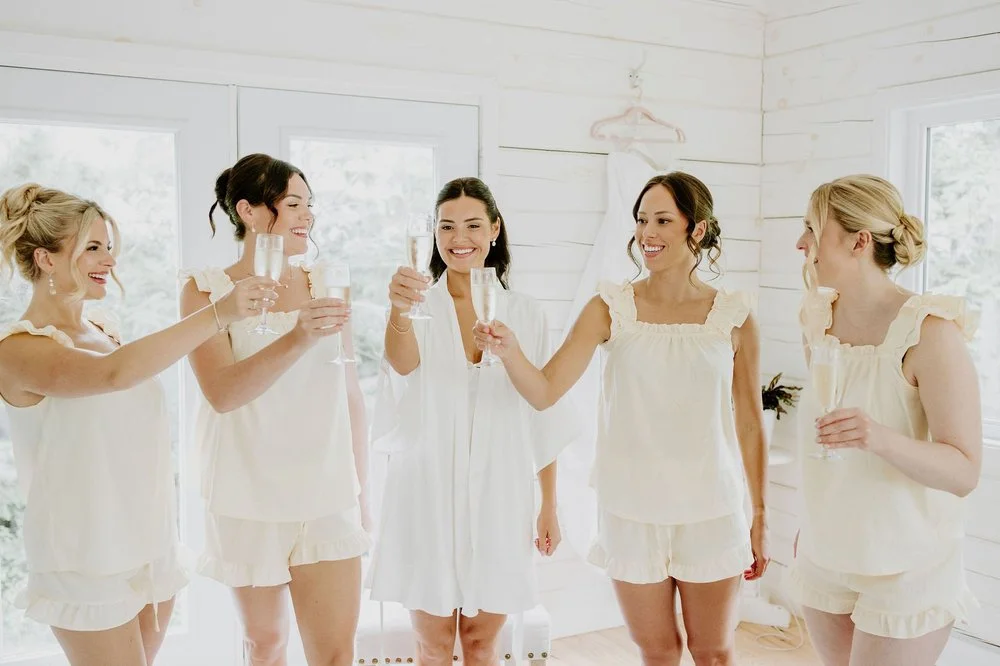 Kylie in a white robe toasting champagne with four bridesmaids in cream pyjama sets