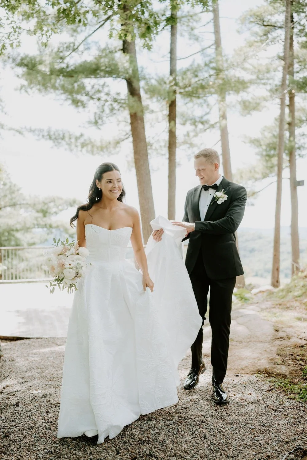 Will carrying Kylie's train as she walks under tall pines with her blush bouquet at Le Belvedere