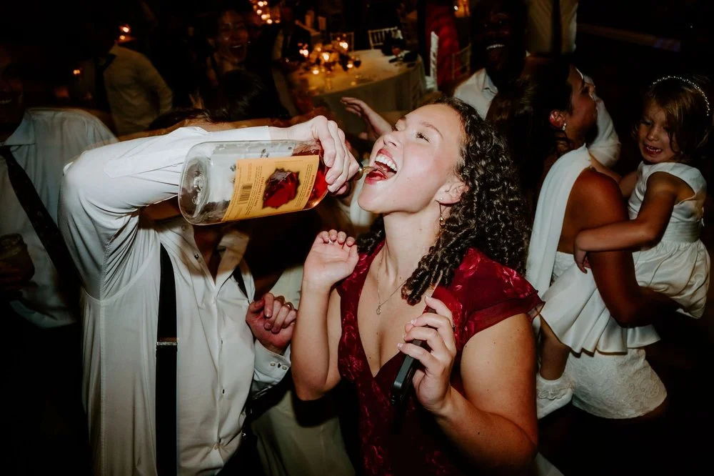 A guest in a red dress laughing as a groomsman pours her a drink straight from the bottle on the dance floor