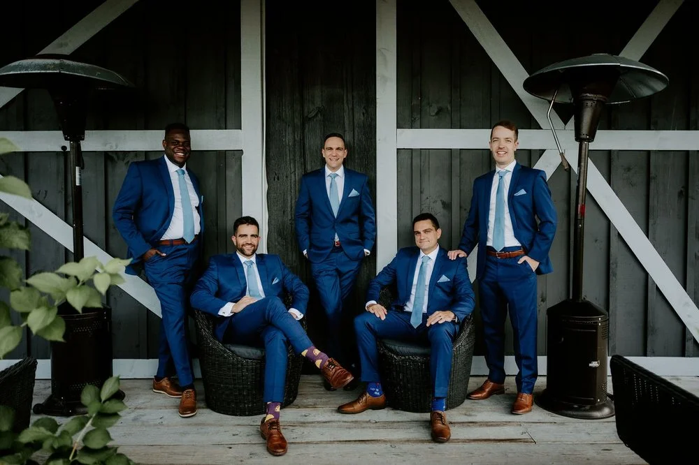Groom and groomsmen in matching blue suits and colourful socks posing in front of a dark barn at Evermore