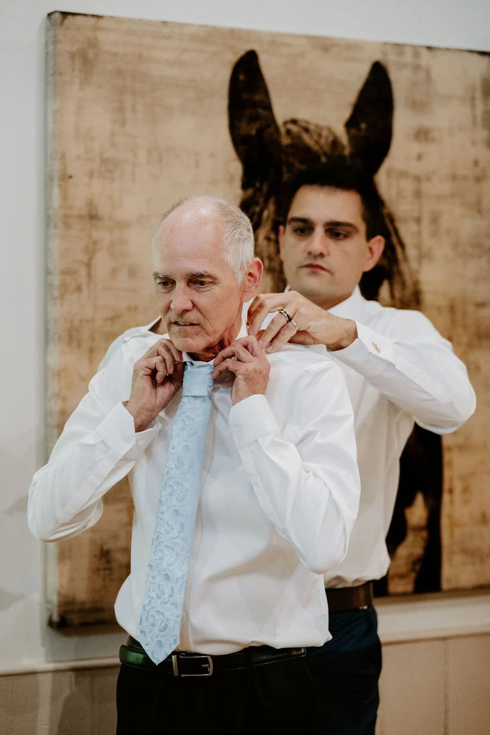 Young man fastening the collar of an older man's shirt and pale blue tie in front of a painting of a horse