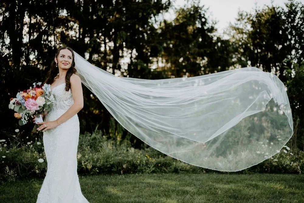 Holly holding her bouquet as her long cathedral veil billows out beside her in the gardens at Evermore
