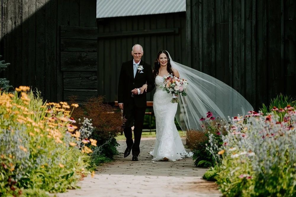 Holly's father walks her down the wildflower-lined garden path at Evermore, her long veil blowing behind her