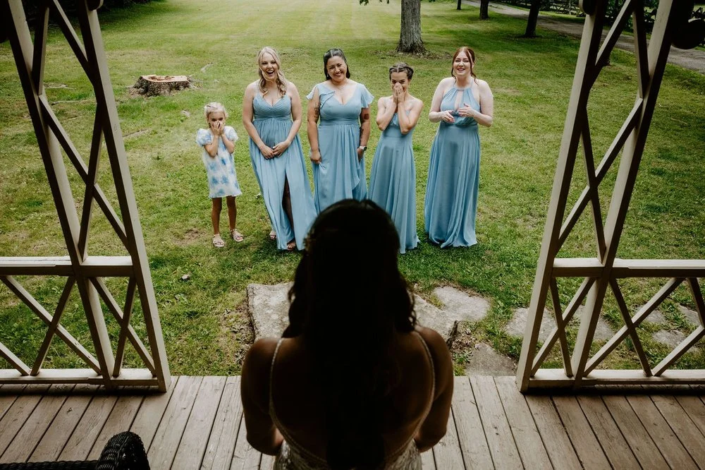 Holly on the porch at Evermore as her bridesmaids in dusty blue and the flower girl react with delight on the lawn