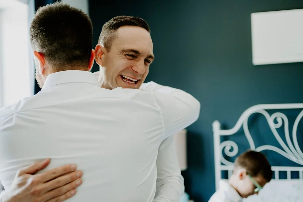 Petar beaming as he hugs a groomsman on the morning of his wedding, with a young boy on a bed in the background