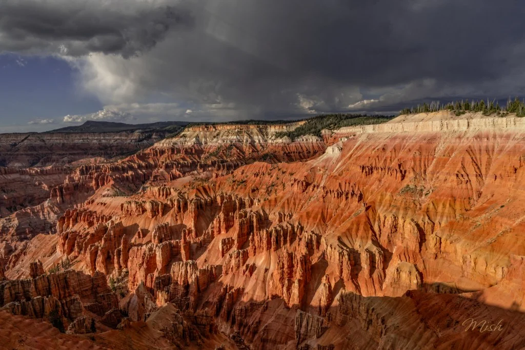 A panoramic view of colorful rock formations and canyons with dramatic shadows and clouds above.