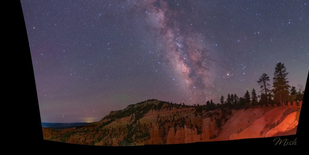 Panoramic view of the Milky Way galaxy against a mountainous landscape with trees and rock formations, showcasing a starry night sky.