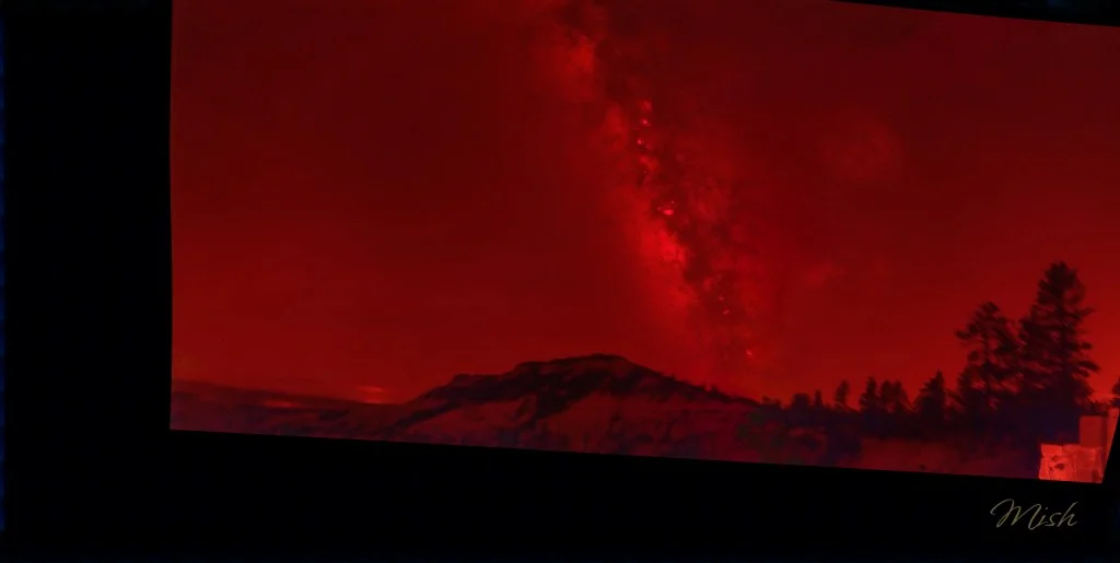 A dramatic landscape scene featuring a silhouette of mountains and trees under a vibrant red sky, showcasing the Milky Way galaxy in striking detail.