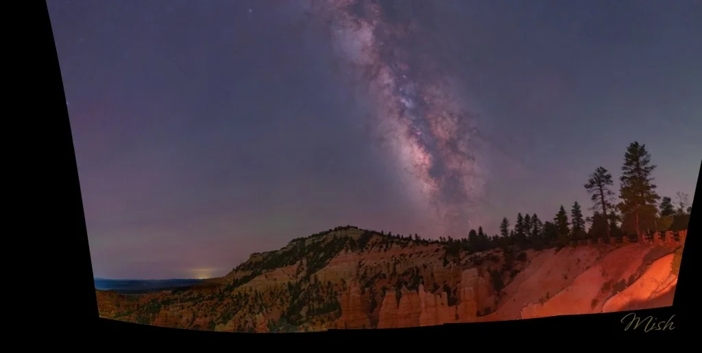 Panoramic view of the Milky Way over rocky terrain with pine trees silhouetted against the night sky.
