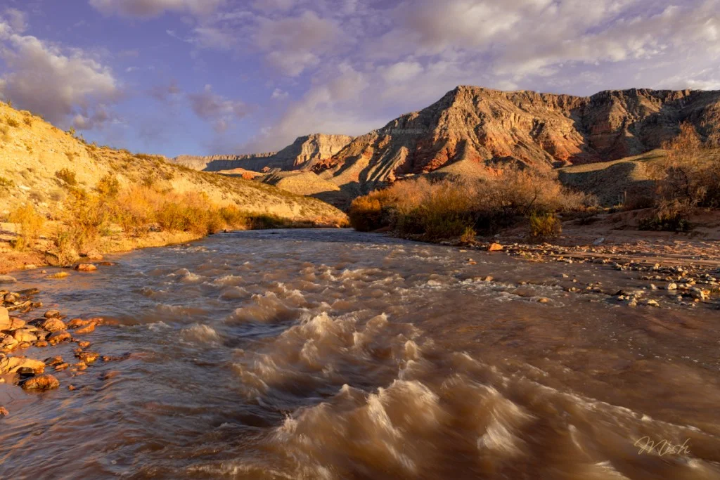 A river with gentle waves flows through a rocky landscape, surrounded by golden autumn foliage and majestic mountains under a partly cloudy sky.