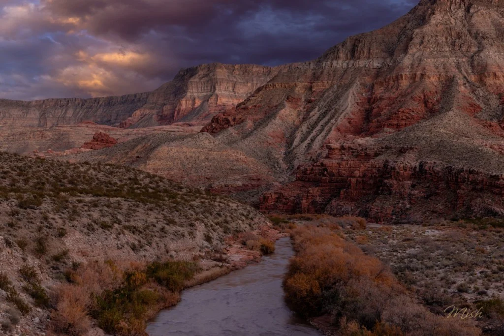 A scenic view of a river winding through rugged, mountainous terrain with colorful rock formations under a dramatic sky.