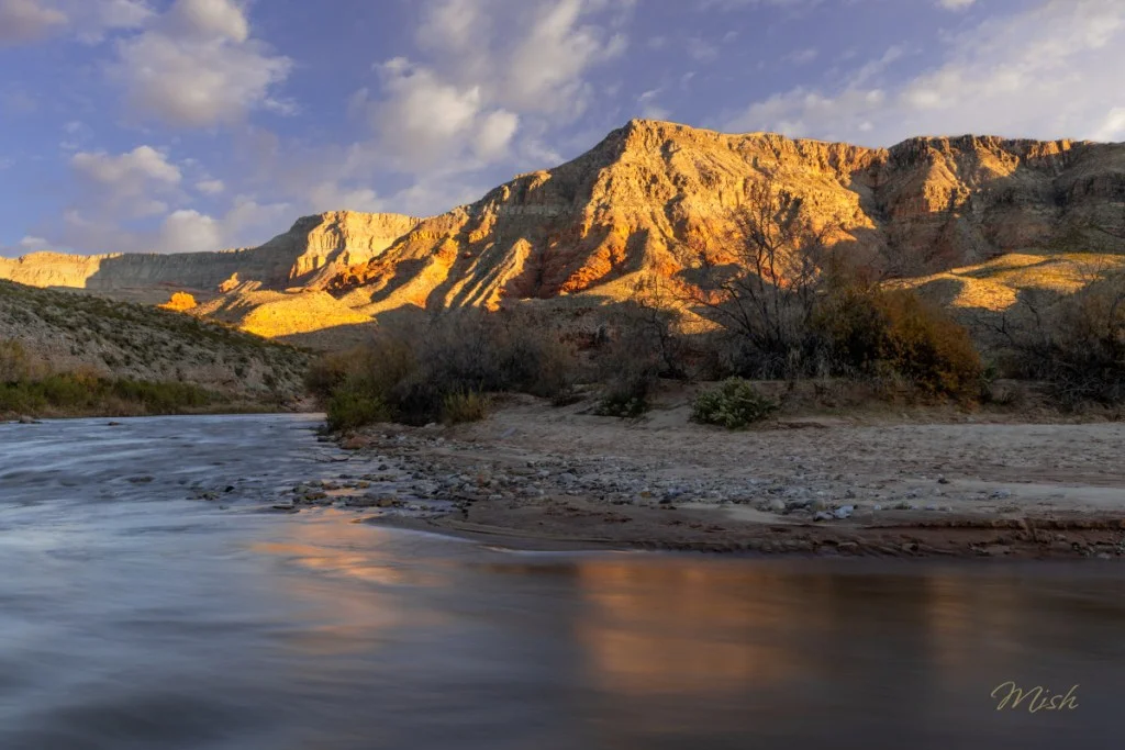 A tranquil river flowing past sandy banks, with rugged, sunlit hills in the background under a partly cloudy sky.