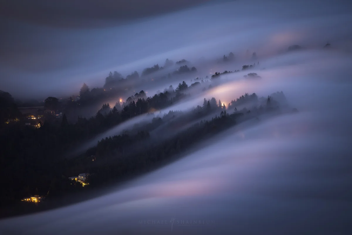 Long exposure of fog flowing over forested hills at blue hour, with layered trees and warm house lights showing atmospheric perspective in a foggy landscape.