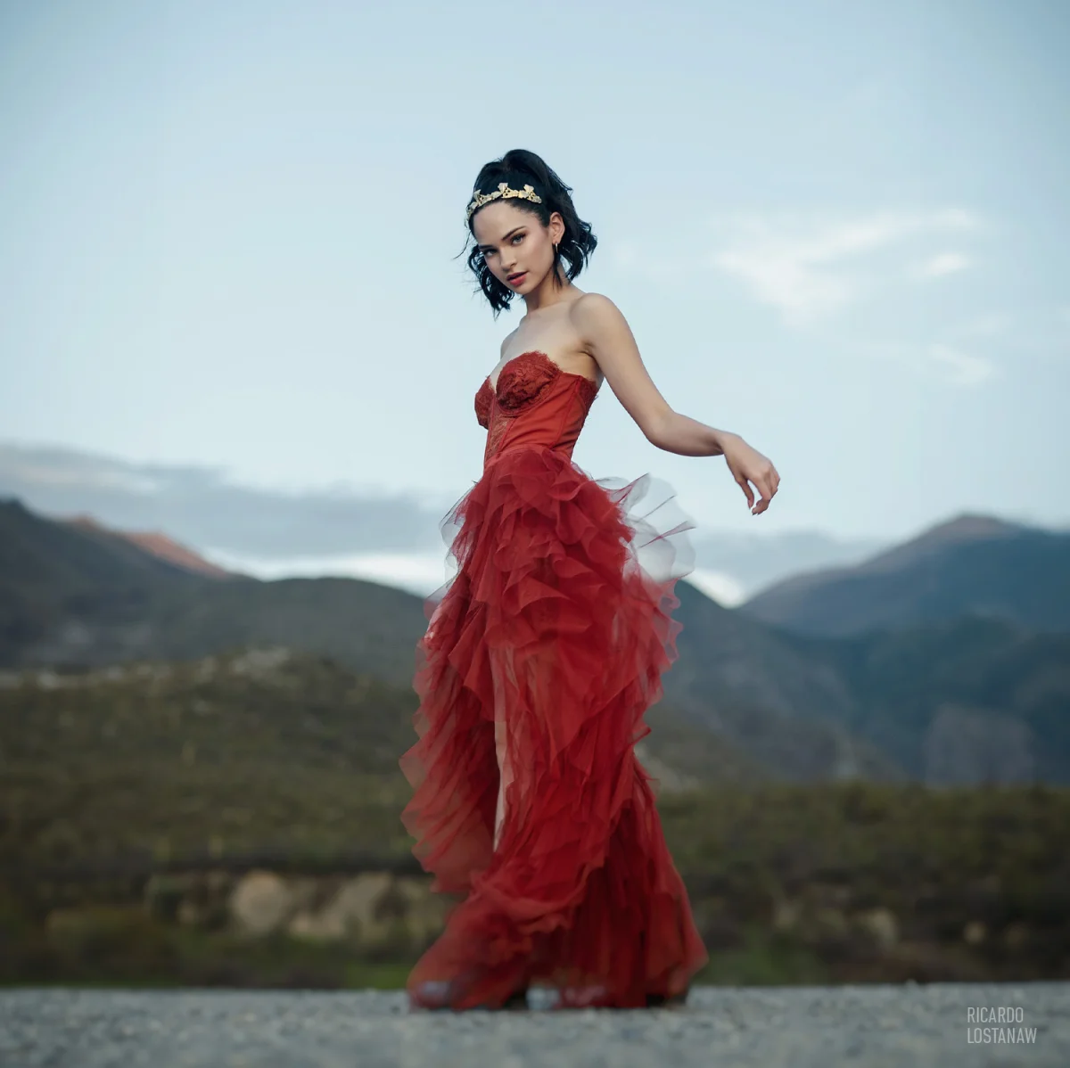 Young woman in a red tulle gown and gold headpiece against a mountain backdrop